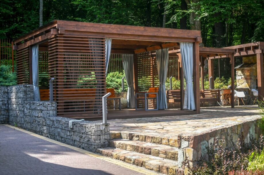 Pergola z ruchomymi deskami drewnianymi, otoczona zielenią i kamienną ścieżką.