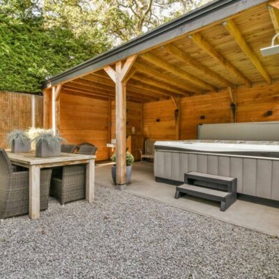 Wiata drewniana z dachem, pod którą znajdują się jacuzzi i stół z krzesłami.